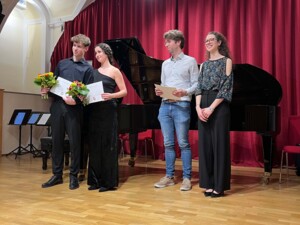 Vier Personen stehen auf der Bühne vor einem Konzertflügel. Zwei Musiker:innen halten Urkunden und Blumensträuße, während zwei weitere Personen daneben stehen. Alle wirken zufrieden und schauen in Richtung Publikum.