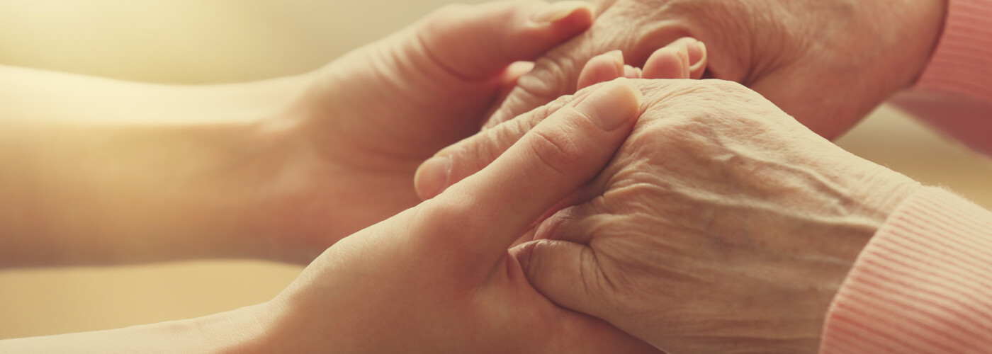 Eine jüngere Person hält sanft die Hände einer älteren Person. Die Szene ist in warmes Licht getaucht und vermittelt Nähe, Trost und gegenseitige Unterstützung.