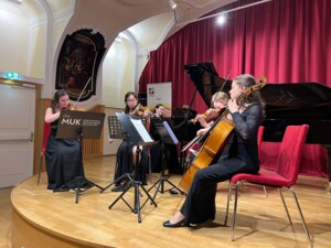 Vier Musiker:innen spielen gemeinsam Streichinstrumente in einem Ensemble. Sie sitzen auf Stühlen mit Notenständern vor sich. Im Hintergrund steht ein Flügel, und ein Schild mit der Aufschrift "die MUK" ist sichtbar.