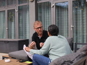 Zwei Teilnehmende sitzen draußen auf einer Couch, sprechen miteinander und betrachten ein Dokument.