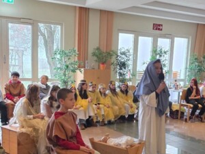 Mehrere Kinder in historischen Kostümen führen ein Krippenspiel auf. Im Vordergrund sitzt eine als Josef verkleidete Person neben einer Holzkrippe mit einer Babypuppe. Daneben steht eine als Maria verkleidete Person mit Mikrofon. Im Hintergrund sitzen weitere Kinder in Engels- und Hirtenkostümen.