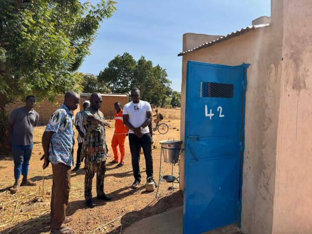 Mehrere Personen stehen im Freien vor einem kleinen Gebäude mit einer auffällig blauen Tür mit der Nummer 42 und betrachten gemeinsam die Situation vor Ort; eine Person zeigt auf den Boden, während andere aufmerksam zuhören. Im Hintergrund sind Bäume, weitere Gebäude und ein Fahrrad zu sehen. Es ist eine Besichtigung in einem ländlichen Umfeld.