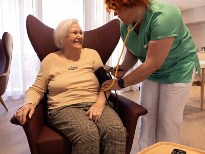 Eine Bewohnerin sitzt auf einem Ohrensessel. Die Pflegerin steht daneben und misst ihren Blutdruck. Die Stimmung ist heiter und entspannt, beide Personen lächeln einander an. Foto: ÖJAB / Erika Mayer.