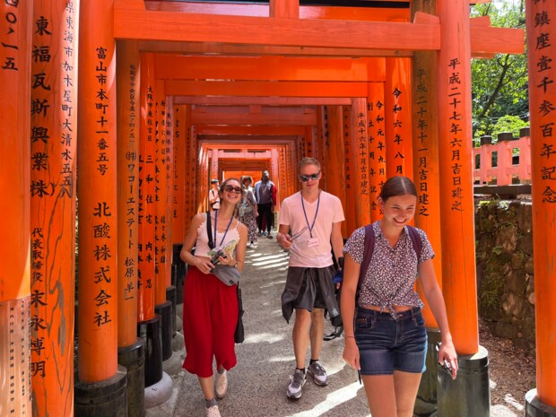 Drei Personen gehen lächelnd durch einen von zahlreichen leuchtend roten Torii-Toren gesäumten Weg eines japanischen Schreins. Die Torii sind mit schwarzen japanischen Schriftzeichen beschriftet und bilden einen langen, tunnelartigen Gang. Sonnenlicht fällt stellenweise durch die Toranlagen und beleuchtet den Weg. Im Hintergrund sind weitere Besucher:innen zu sehen.