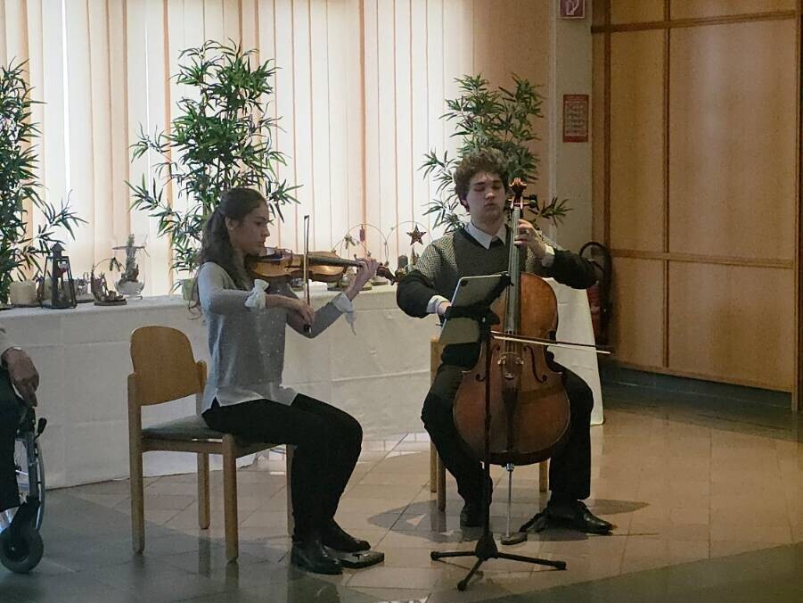 Zwei junge Musiker:innen sitzen nebeneinander in einem festlich geschmückten Raum und spielen ein Adventkonzert. Links spielt eine Person Violine, rechts eine Person Cello. Beide sind konzentriert bei der Darbietung. Im Hintergrund stehen Pflanzen und eine weiße Tafel mit weihnachtlicher Dekoration.