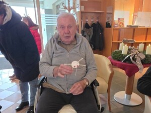 Ein älterer Mann im Rollstuhl hält stolz ein selbstgebasteltes Geschenk in der Hand. Neben ihm ein Adventskranz auf einem Tisch, im Hintergrund weihnachtliche Dekoration.