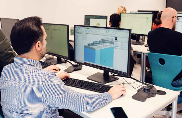 Ein Kursteilnehmer sitzt in einem Computerraum vor zwei Monitoren. Auf dem Bildschirm ist ein digitales 3D-Modell mit technischen Bauteilen zu sehen. Weitere Kursteilnehmende arbeiten im Hintergrund an Computerarbeitsplätzen. Foto: www.inframe.at