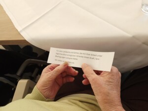 Eine Person hält einen Zettel mit einer vorgedruckten Fürbitte in der Hand. Auf dem Tisch liegt eine herbstliche Dekoration mit Kürbis, Kastanien und Blumen.