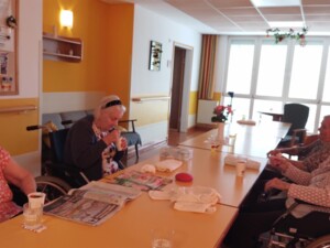 Mehrere ältere Bewohner:innen sitzen in einem Gemeinschaftsraum an einem langen Tisch. Einige halten Becher mit Sommerbowle, auf dem Tisch liegen Zeitungen und Gläser.