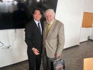 Zwei Personen stehen nebeneinander in einem Innenraum vor einem großen Bildschirm und blicken lächelnd in die Kamera. Links trägt eine jüngere Person einen dunklen Anzug mit Krawatte, rechts steht eine ältere Person mit weißem Haar in einem karierten Sakko und gemusterter Krawatte. Die ältere Person hält eine Geschenktasche in der Hand. Der Raum ist ein Seminar- oder Veranstaltungsraum mit Garderobenhaken und einem Tisch.