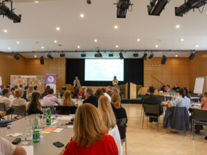 Großer Veranstaltungssaal mit vielen Teilnehmenden an Tischen, die auf eine Bühne mit Präsentationsfolie und Vortragenden blicken.