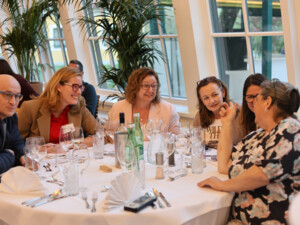 Mehrere Teilnehmende sitzen an einem gedeckten Tisch, lachen und unterhalten sich in entspannter Atmosphäre.