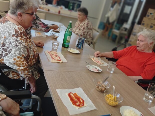 Mehrere Senior:innen sitzen um einen großen Tisch und bereiten gemeinsam Pizzen zu. Auf den Teigstücken ist bereits Tomatensauce verteilt. In Schälchen stehen Zutaten wie Mais, Käse und Pilze bereit. Die Stimmung wirkt lebendig und gesellig.