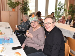 Eine ältere Frau mit selbstgebasteltem, silbernem Faschingshut sitzt lächelnd neben einem Mann mit Brille. Beide sitzen an einem Tisch mit Krapfen und Getränken. Im Hintergrund sind weitere Bewohner:innen an festlich gedeckten Tischen zu sehen.