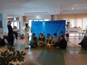 Ein:e Jugendliche:r in weißem Kostüm und mit Flügeln steht mit Mikrofon vor einer Gruppe sitzender Hirten-Darsteller:innen. Ein:e weitere:r Schüler:in hebt eine Lampe hoch. Die Szene spielt vor einem Kulissenbild mit Nachthimmel.