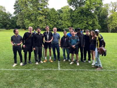 Gruppenbild eines Teams in schwarzer Sportkleidung, das einen Pokal hält. Die Gruppe steht auf einer Wiese, im Hintergrund sind weitere Personen und Bäume sichtbar.