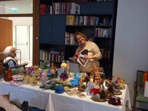 Eine Person steht hinter einem Tisch voller bunter, selbstgemachter Weihnachtsdekorationen wie Kerzen, Figuren, kleinen Geschenken und Naturmaterialien. Im Hintergrund befindet sich ein großes Bücherregal. Die Szene vermittelt eine kreative und festliche Marktatmosphäre im Innenraum.