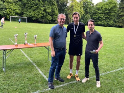 Drei Personen stehen vor einem Tisch mit Pokalen, eine davon trägt eine Medaille um den Hals. Im Hintergrund ist ein Fußballtor zu sehen.