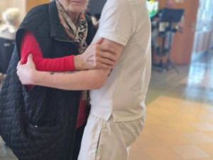 Eine ältere Frau mit weißem Haar, rotem Pullover und schwarzer Weste tanzt mit einer jungen Pflegerin in weißer Kleidung. Beide umarmen sich und lächeln.