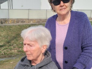 Eine ältere Bewohnerin sitzt in einem Rollstuhl, hinter ihr steht eine Begleitperson mit Sonnenbrille und violetter Strickjacke. Beide sind draußen auf einem Weg bei sonnigem Himmel unterwegs.