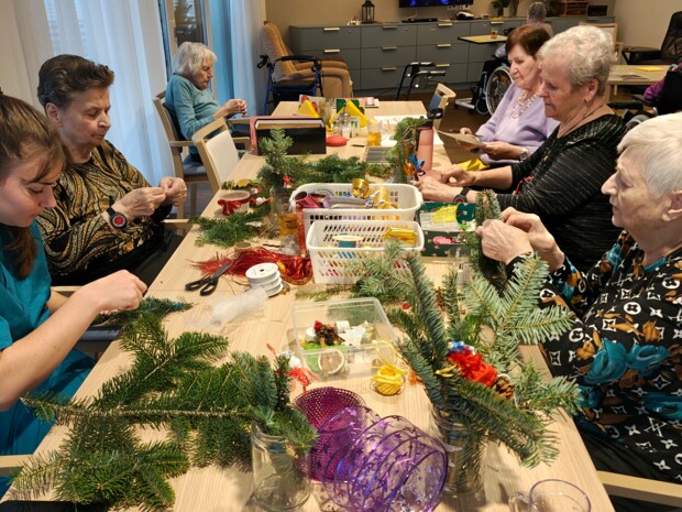 Mehrere Bewohner:innen sitzen gemeinsam mit einer Betreuungsperson an einem Tisch und basteln Weihnachtsdekoration. Auf dem Tisch liegen Tannenzweige, Schleifen, Scheren und kleine Dekorationselemente. Die Teilnehmenden arbeiten konzentriert und tauschen sich dabei aus. Die Atmosphäre ist ruhig und kreativ.