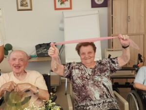 Eine Bewohnerin hält ein rosa Tuch mit ausgestreckten Armen hoch und lächelt. Neben ihr sitzen weitere Bewohner:innen, eine Person hält ein grünes Tuch, eine andere sitzt im Rollstuhl mit einer Flasche in der Hand.