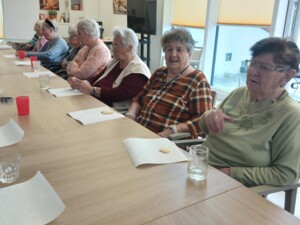 Mehrere Senior:innen sitzen nebeneinander an einem langen Tisch mit Gläsern und Servietten. Jede Person hat ein Keksstück vor sich liegen.