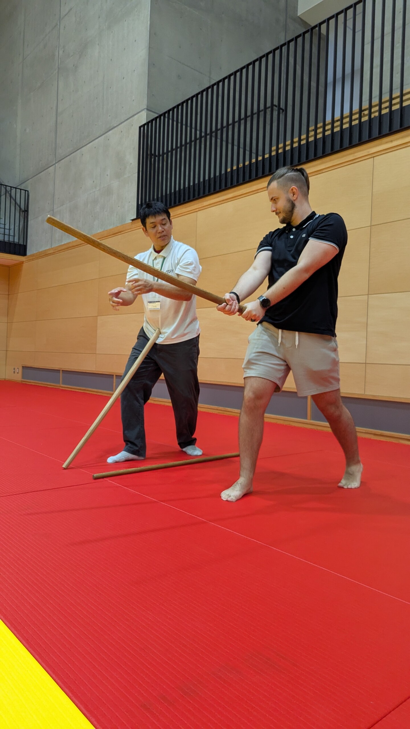 A Japanese instructor demonstrates the correct posture in Aikido with a wooden staff to a barefoot Austrian participant training on a red mat.