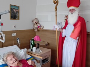 Eine ältere Person liegt in einem Pflegebett und lacht, während der Nikolaus danebensteht und ein rotes Geschenk bereithält. Auf dem Nachttisch stehen Getränke und persönliche Gegenstände.