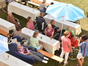 Blick von oben auf lange Tische im Garten. Bewohner:innen, viele im Rollstuhl, sitzen unter blauen Sonnenschirmen. Einige unterhalten sich angeregt, andere schauen in Richtung Musik.
