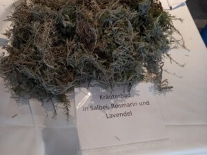 Ein weiß gedeckter Tisch mit einem großen Bündel getrockneter Kräuter wie Lavendel, Rosmarin und Salbei. Darunter ein Schild mit der Beschriftung "Kräuterbad in Salbei, Rosmarin und Lavendel". Eine Station für die Sinne.