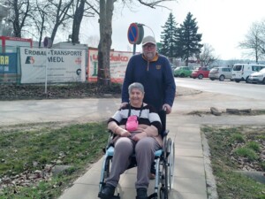 Eine Bewohnerin sitzt lächelnd in einem Rollstuhl auf einem Gehweg, hinter ihr steht eine Begleitperson und schiebt den Rollstuhl. Im Hintergrund sind eine Straße, parkende Autos und Baustellenzäune zu sehen.