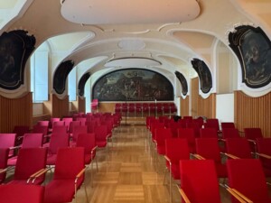 Ein leerer Konzertsaal mit Reihen roter Stühle und einer Bühne im Hintergrund. Die Decke ist kunstvoll gestaltet, und an den Wänden hängen große Gemälde. Der Raum wirkt ruhig und bereit für ein Publikum.