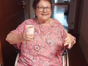 Eine ältere Bewohnerin sitzt lächelnd in einem Rollstuhl in einem Flur und hält einen Becher mit Sommerbowle in der Hand. Im Hintergrund ist ein heller Wohnbereich zu sehen.