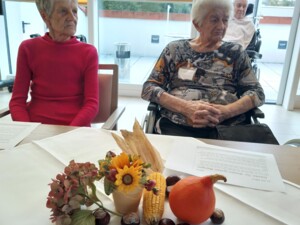 Zwei ältere Personen sitzen an einem Tisch mit einer herbstlichen Tischdekoration aus Sonnenblumen, Kürbis, Maiskolben, Kastanien und Hortensien. Auf dem Tisch liegen auch Fürbitt-Texte.
