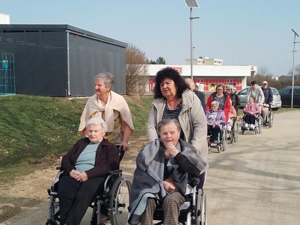 Mehrere Bewohner:innen in Rollstühlen sind mit Begleitpersonen auf einem Gehweg im Freien unterwegs. Die Gruppe bewegt sich bei sonnigem Wetter Richtung Stadtstraße, im Hintergrund stehen Autos und Bäume.