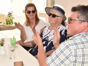 Drei Personen sitzen an einem Tisch im Freien und klatschen. Eine Seniorin trägt einen Sommerhut, daneben zwei Angehörige mit Sonnenbrillen. Auf dem Tisch stehen Gläser und eine kleine Blumenvase.