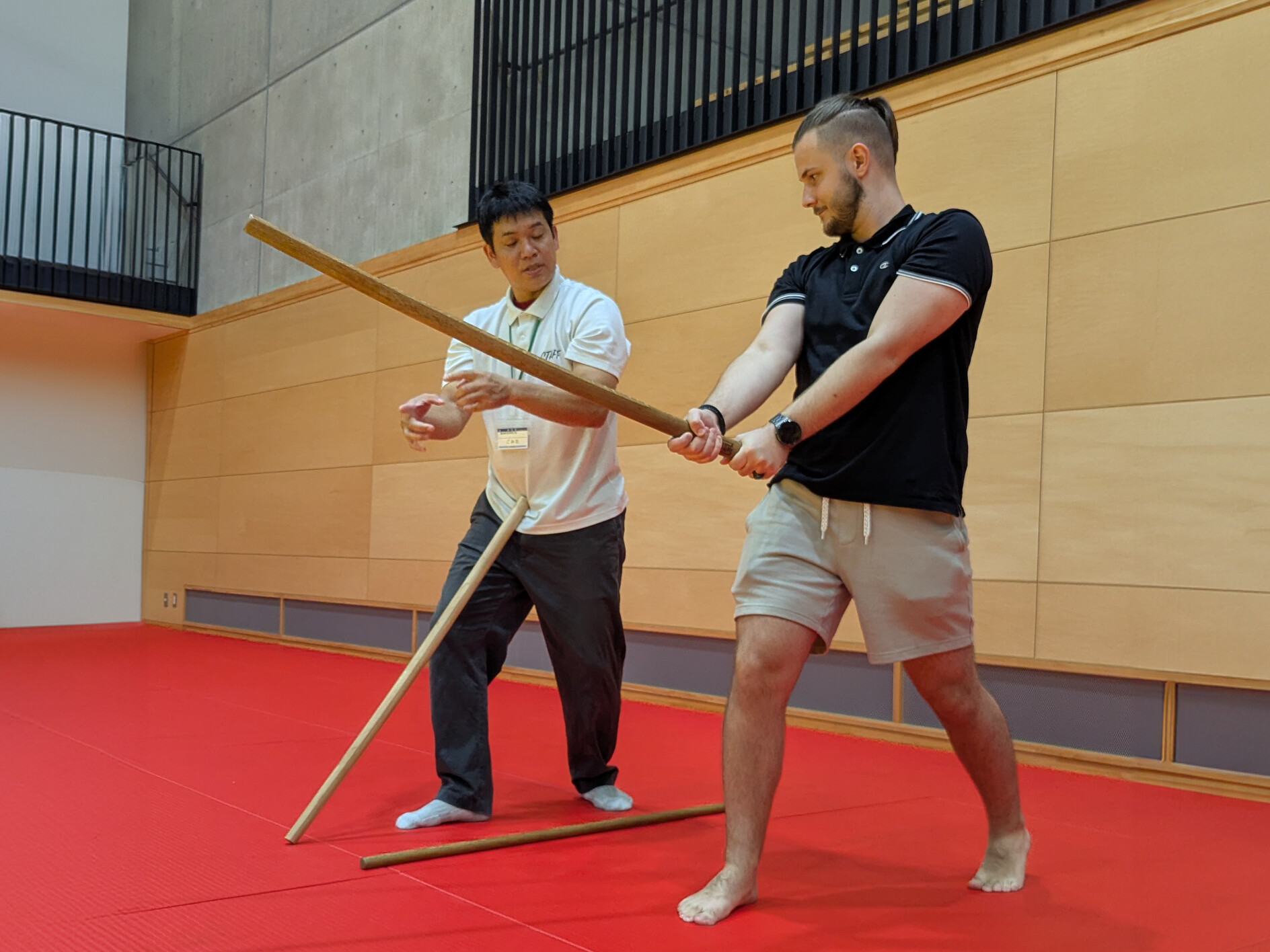 Eine japanische Lehrperson zeigt einem barfuß trainierenden österreichischen Teilnehmer auf roter Matte die korrekte Haltung beim Aikido mit einem Holzstab.