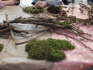 Eine ältere Person sitzt an einem Tisch und arrangiert Moosstücke für eine herbstliche Bastelarbeit. Auf dem Tisch liegen auch Kastanien, Nüsse und Zweige. Im Hintergrund sind weitere Personen und Bastelmaterialien zu sehen.