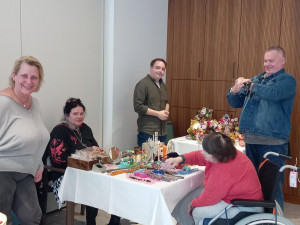 Eine Gruppe von fünf Personen, darunter eine Rollstuhlnutzerin, steht und sitzt um zwei Tische mit handgefertigtem Schmuck und Osterartikeln.