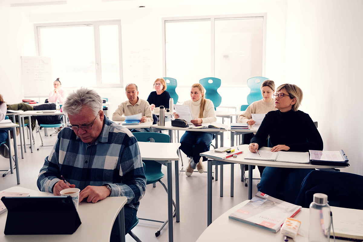 Mehrere Kursteilnehmende sitzen in einem hellen Unterrichtsraum an einzelnen Tischen. Einige lesen oder halten Arbeitsblätter, eine Person im Vordergrund schreibt neben einem Tablet Notizen. Foto: www.inframe.at