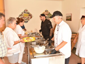 Zwei Köche und weitere Mitarbeitende geben an einer Buffetstation Speisen aus. Gäste stehen mit Tellern in der Hand in der Schlange. Auf dem Buffet liegen verschiedene Fleisch- und Gemüsegerichte.