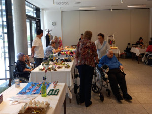 Weitwinkelaufnahme des Veranstaltungsraums mit mehreren Tischen voller Osterdekorationen und vielen Besucher:innen, darunter auch Rollstuhlnutzer:innen.