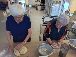 Zwei Senior:innen bearbeiten Hefeteig - eine Person schneidet ihn, die andere rollt ihn zu einem Strang. Mehl und Rührschüsseln stehen bereit.