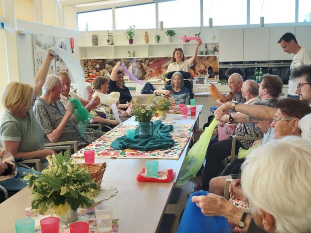 In einem hellen Gemeinschaftsraum sitzen Bewohner:innen rund um einen langen Tisch. Gemeinsam mit Schüler:innen der SOB Güssing bewegen sie farbige Tücher im Sitzen. Auf dem Tisch stehen Blumen, Becher und Klangschalen.