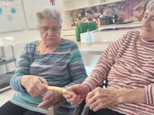 Zwei ältere Bewohnerinnen sitzen nebeneinander und halten gemeinsam einen gebackenen Fastenbeugel, den sie vorsichtig auseinanderziehen. Hinter ihnen befindet sich eine offene Küchenzeile mit Flaschen, Pflanzen und dekorativen Elementen.