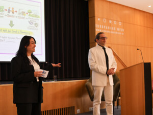 Zwei Moderator:innen stehen auf der Bühne, präsentieren Inhalte und führen durch das Programm vor Publikum.