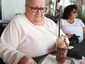 Eine Seniorin sitzt an einem Tisch und betrachtet konzentriert ein großes Glas mit Eiskaffee, Sahne und Schokodekor. Im Hintergrund sitzt eine weitere Person mit Sonnenbrille.