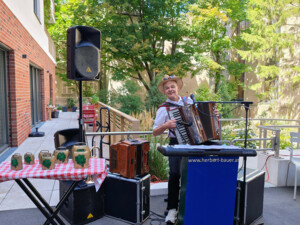 Ein Musiker steht vor einem Mikrofon im Gartenbereich und spielt auf einer Steirischen Harmonika. Er trägt einen Strohhut und lächelt. Um ihn herum stehen Lautsprecher, weitere Instrumente sowie ein Tisch mit Dekoration. Im Hintergrund ist üppiges Grün zu sehen.