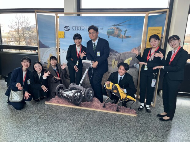 Acht Personen in formeller Kleidung posieren gemeinsam vor einem Ausstellungsstand der CTBTO mit technischem Hintergrundmotiv. In der Mitte stehen und knien mehrere Teilnehmende rund um zwei Robotik-Modelle: ein fahrbares Gerät mit Rädern und ein gelber Laufroboter. Alle lächeln in die Kamera, einige heben die Hände in einer präsentierenden Geste. Im Hintergrund zeigt die Standwand eine Einsatzszene mit Hubschrauber und Fachkräften in Schutzanzügen, was den technisch-wissenschaftlichen Kontext des Auftritts unterstreicht.